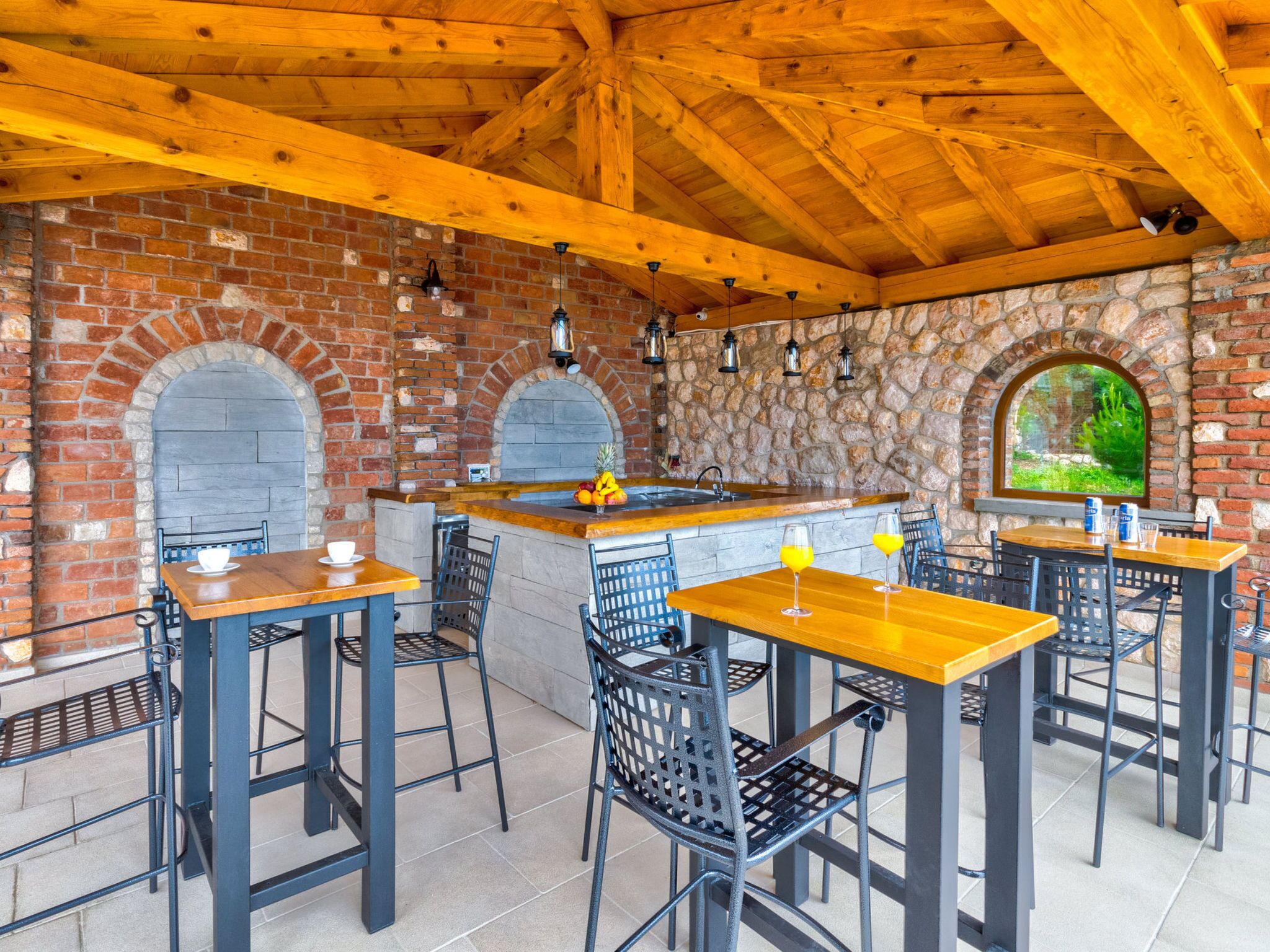 Une terrasse extérieure avec des murs en briques, des arcades en pierre, des poutres en bois, ainsi que des chaises et des tables en métal.