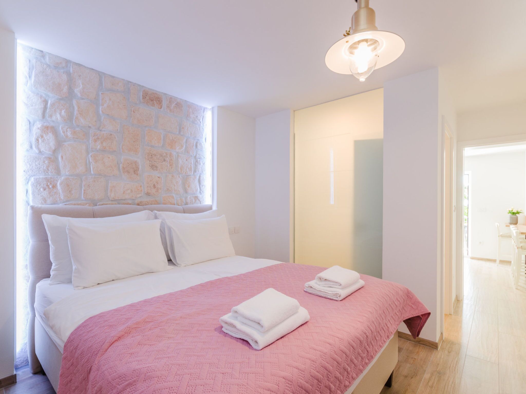 Un dormitorio con pared de piedra, cama con ropa de cama blanca, colcha rosa y lámpara colgante.