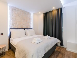 Ein modernes Schlafzimmer mit einer Wand aus Stein, einem großen Bett mit weißer Bettwäsche und Blick ins Freie durch dunkle Vorhänge.