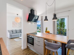 Eine moderne Küche mit Einbauofen, ein Essbereich mit Holztisch und grauen Stühlen, ein graues Sofa im Wohnbereich und ein großes Fenster mit Blick ins Grüne.