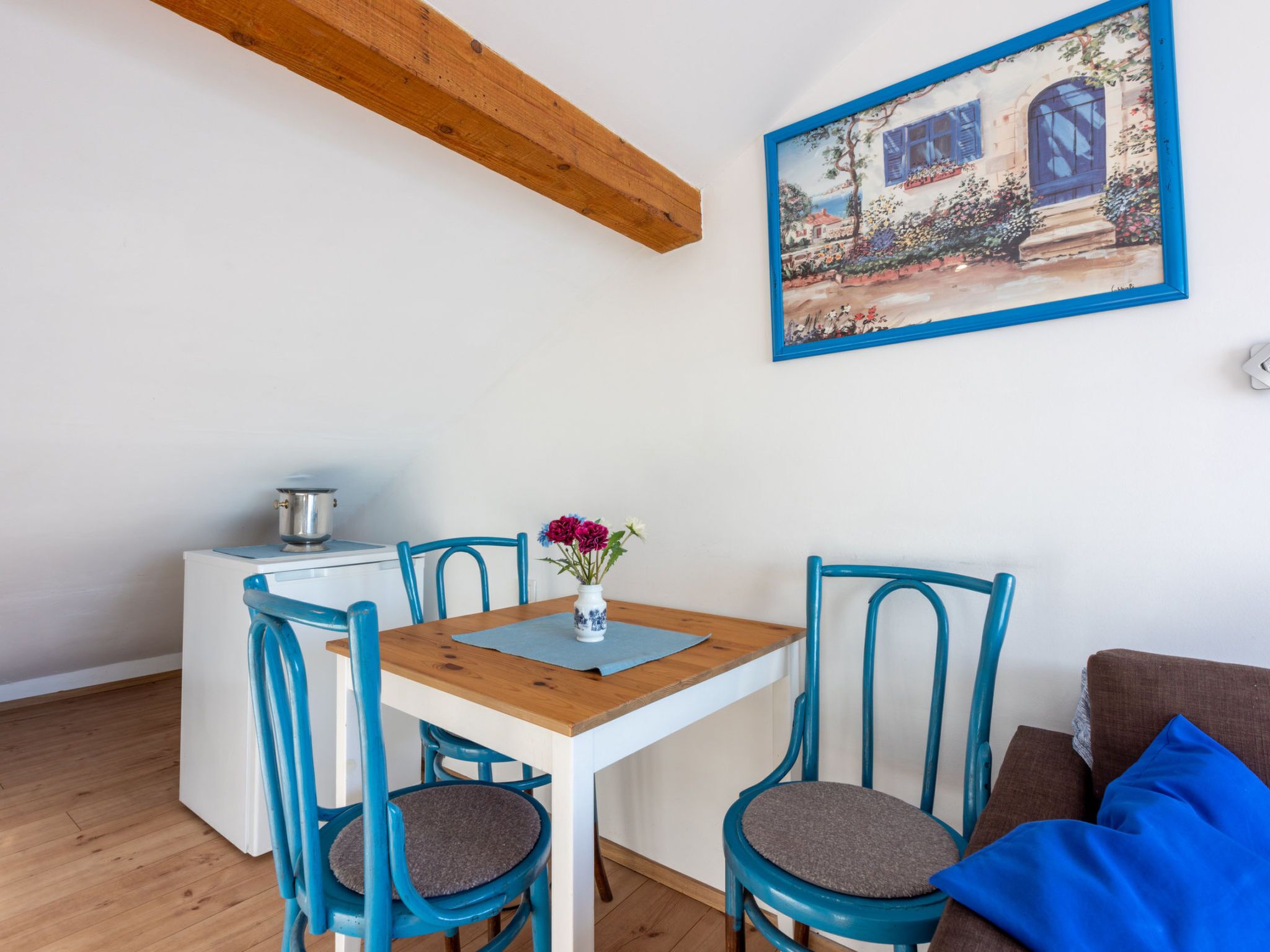 A dining area with a wooden table, blue chairs, a small vase with flowers, and a painting on the wall.