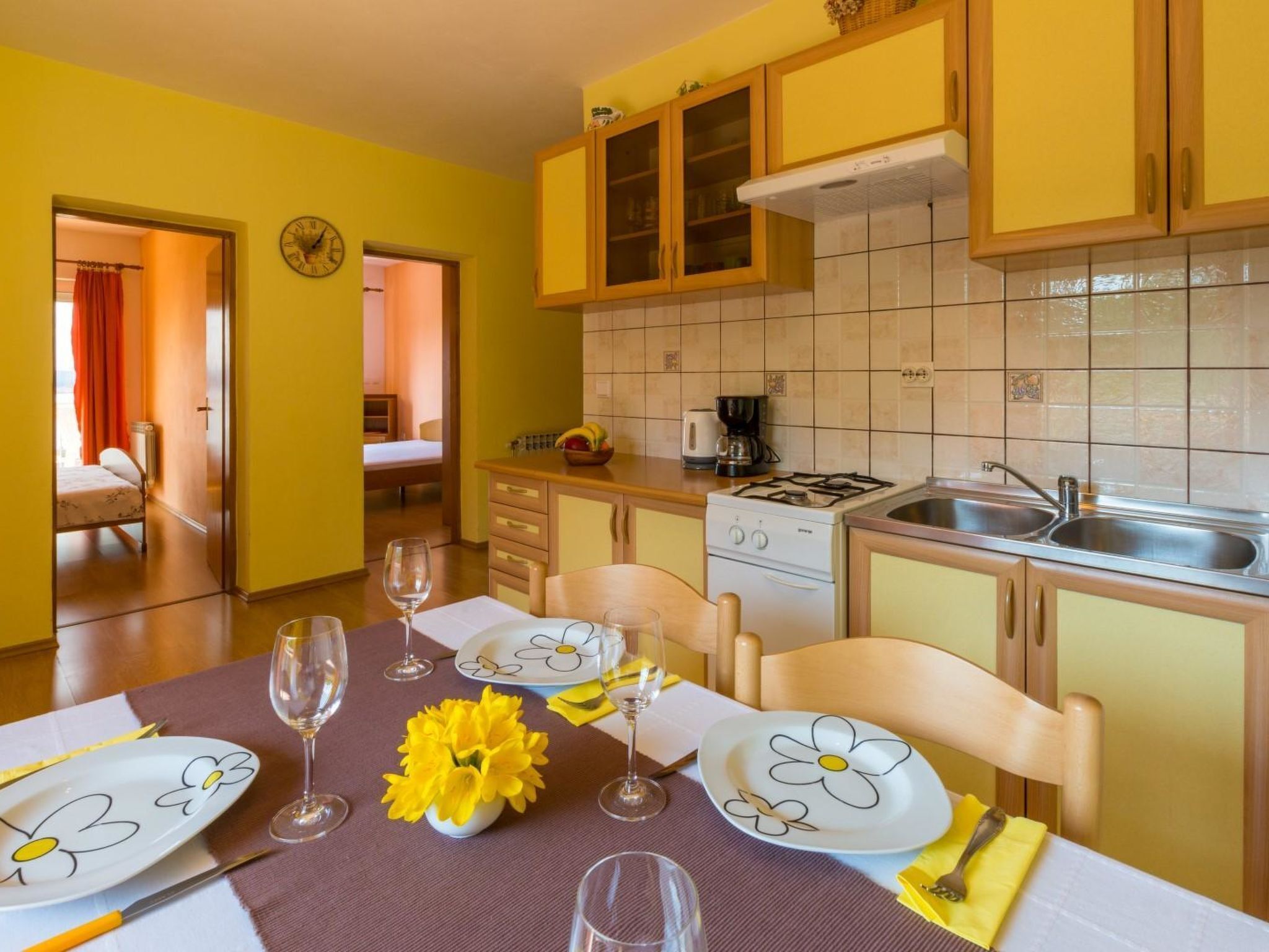 A dining area with a table set for two, a kitchen with yellow cabinets, a stove, and a sink.