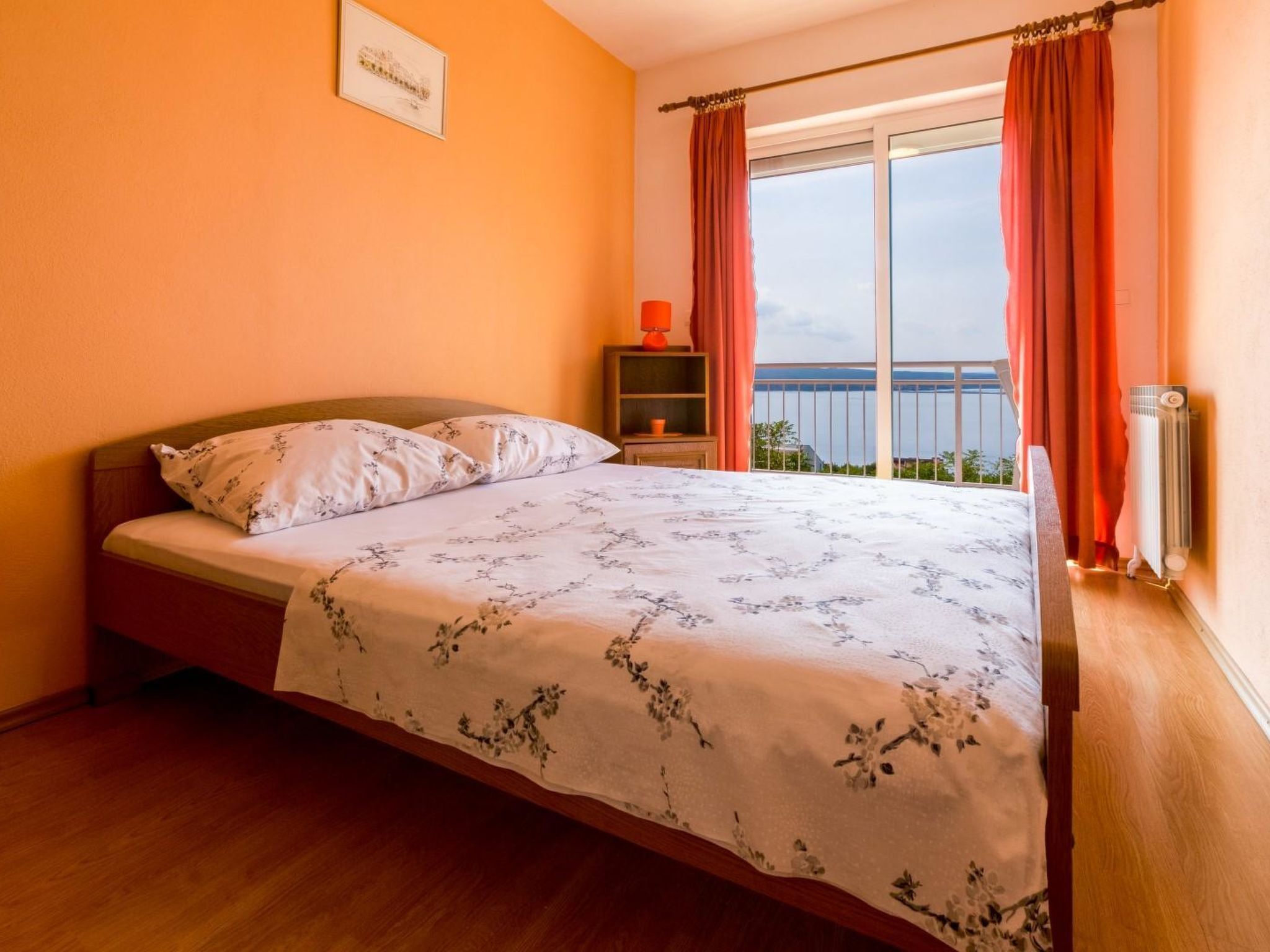 A bedroom with a bed, a nightstand, a window with curtains, and a view of the sea.
