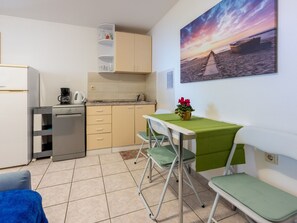 Eine kompakte Küche mit Kaffeemaschine, Wasserkocher und Geschirrspüler. Ein Esstisch mit grüner Tischdecke und zwei Stühle laden zum Verweilen ein. An der Wand hängt ein Bild mit einer Strandansicht.