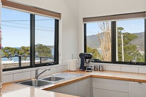 A modern kitchen with a built-in sink, a coffee machine, and large windows offering a view of the sea and trees. - Coastal 2BR Escape, Steps to Beach, Dining and Bar (Eaglehawk Neck)
