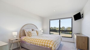 Quarto com cama, mesa de cabeceira, luminária, cômoda e televisão montada na parede.