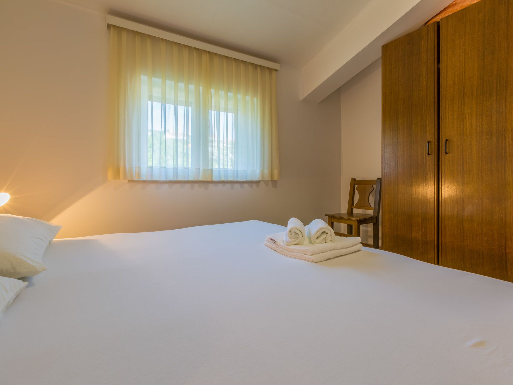 Ett hotellrum med en säng, ett fönster med gardiner, en garderob i trä och en stol.