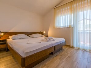A hotel room with a wooden bed, white linens, a nightstand, and a window with sheer curtains. - IRIS 4 / HR-02362-04 by Interhome (Crikvenica)