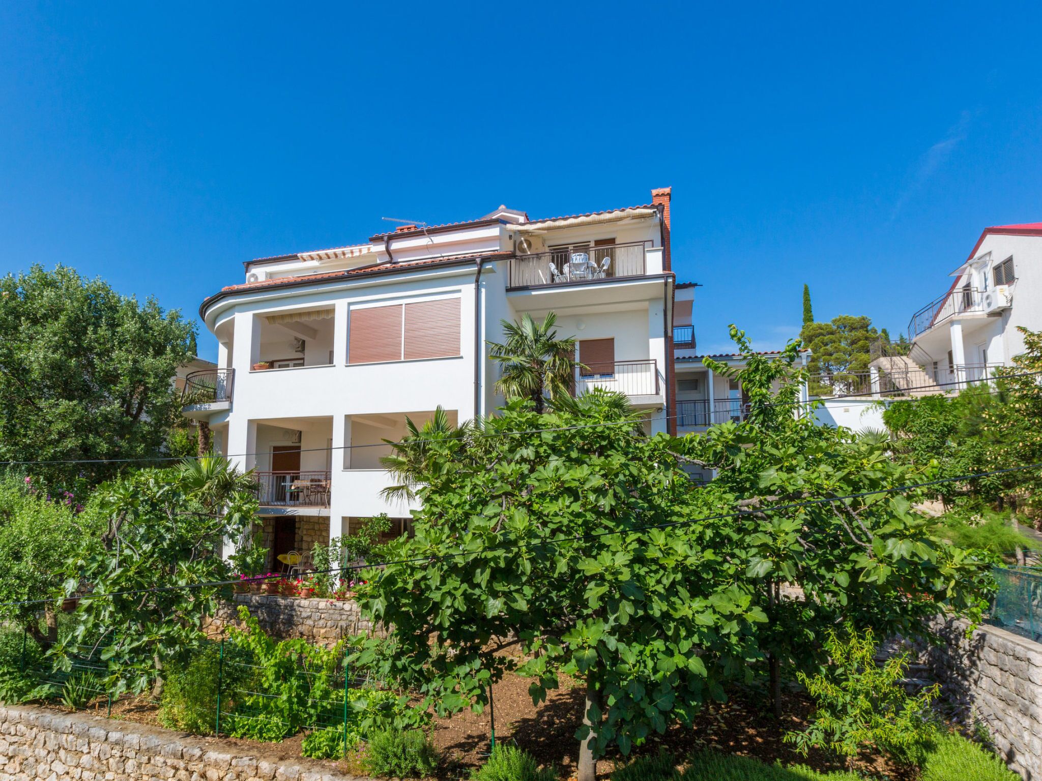 Um edifício residencial de dois andares com varanda, rodeado por vegetação exuberante e um céu azul límpido.