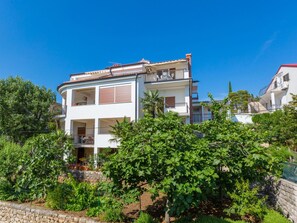 Ein zweistöckiges Wohngebäude mit Balkon, umgeben von üppiger Vegetation und blauem Himmel.