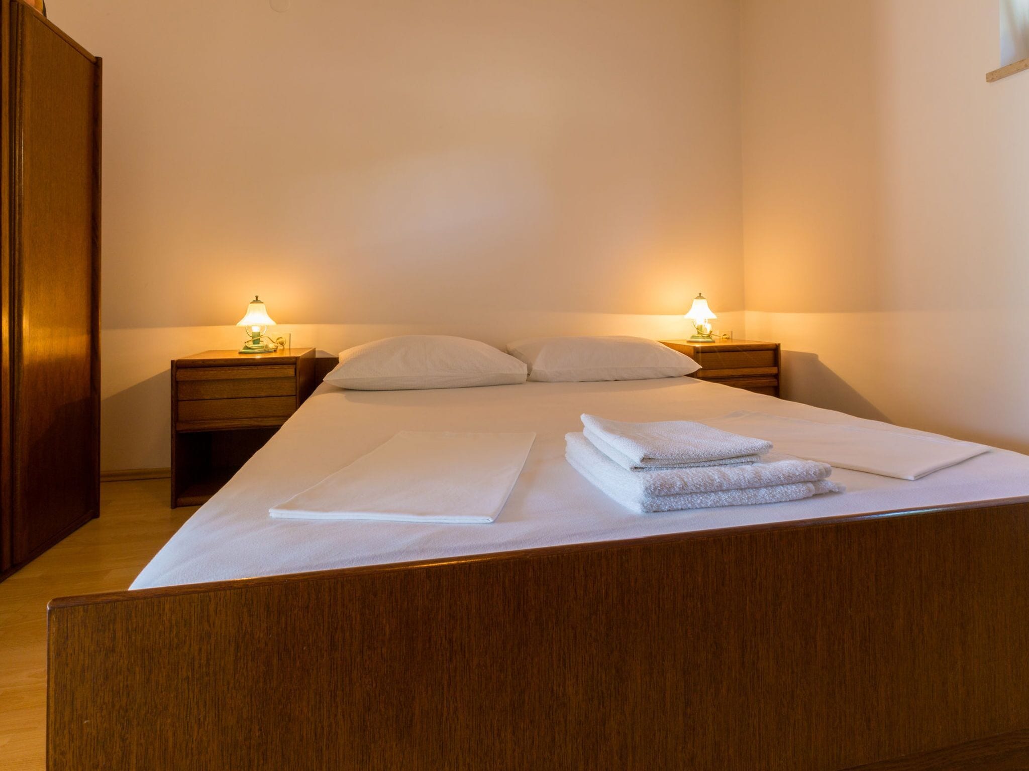 A neatly made bed with white linens and towels, two bedside tables with lamps, and a wooden wardrobe.