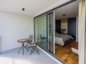 Un balcón con una mesa y sillas, una puerta corrediza de vidrio que da a un dormitorio con una cama y una mesita de noche.