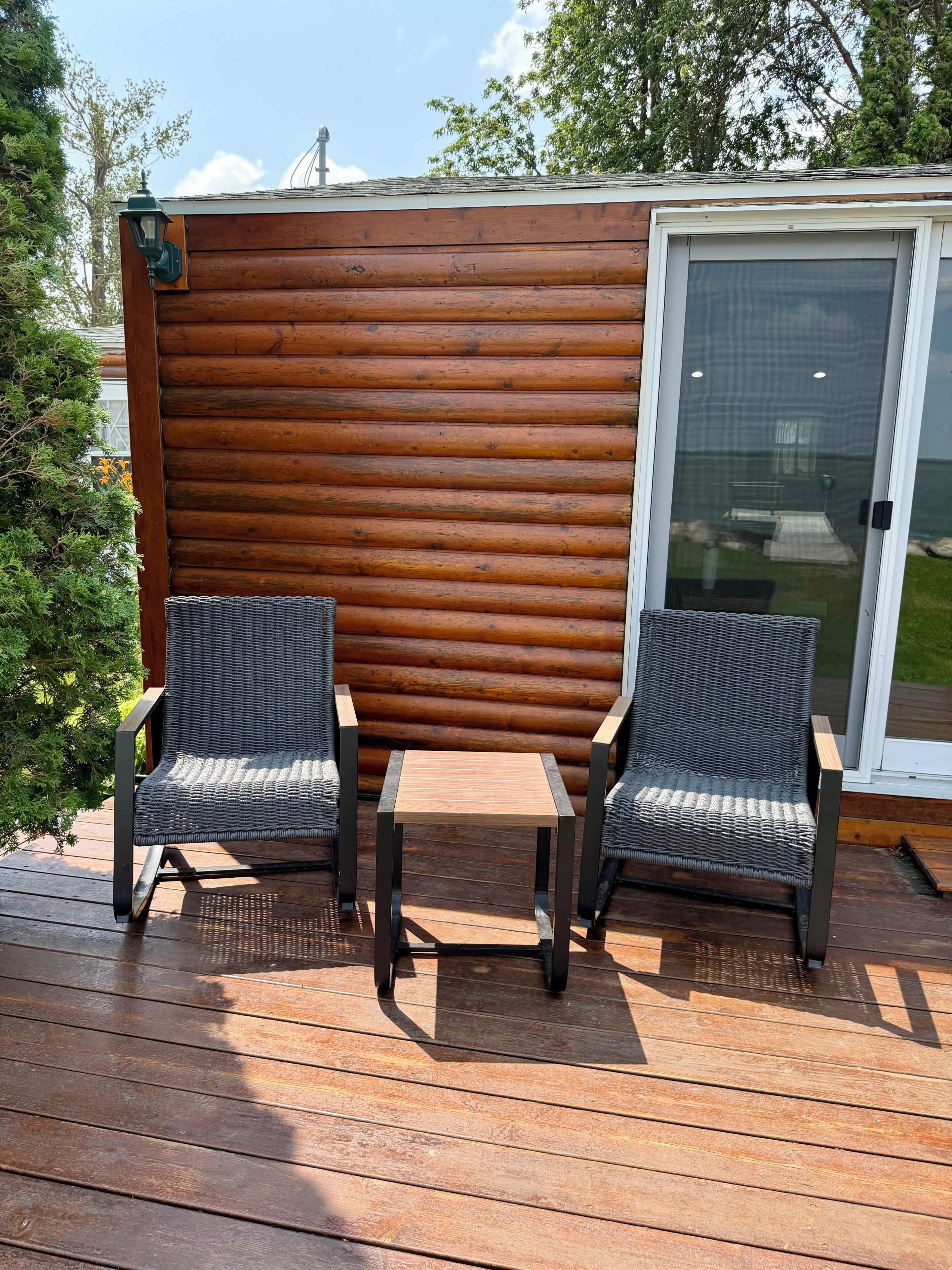 A wooden deck with two wicker chairs and a small table, a sliding glass door, and a backdrop of a wooden fence.