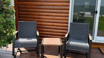Terraza de madera con dos sillas de mimbre y una mesita, un ventanal corredero y un seto de madera al fondo.