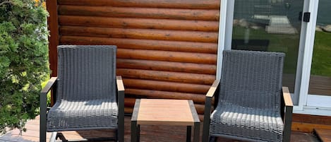 A wooden deck with two wicker chairs and a small table, a sliding glass door, and a backdrop of a wooden fence.