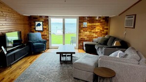 A living room with a television, seating, a coffee table, and a view through a glass door. - Relaxing 3BR Cabin on Blue Dog Lake I Screened Porch I Fireplace I Lakefront (Waubay)
