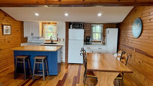 A compact kitchen with wooden walls, a central island, bar stools, a dining table, and a refrigerator. - Relaxing 3BR Cabin on Blue Dog Lake I Screened Porch I Fireplace I Lakefront (Waubay)
