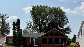 A wooden cabin with a deck leading to a pond, surrounded by rocks and greenery. - Relaxing 3BR Cabin on Blue Dog Lake I Screened Porch I Fireplace I Lakefront (Waubay)
