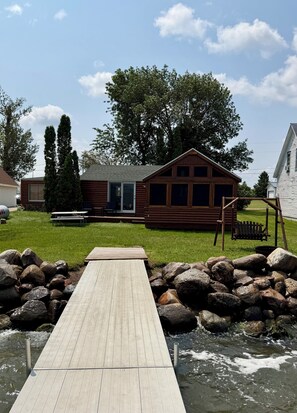 A wooden cabin with a deck leading to a pond, surrounded by rocks and greenery. - Relaxing 3BR Cabin on Blue Dog Lake I Screened Porch I Fireplace I Lakefront (Waubay)