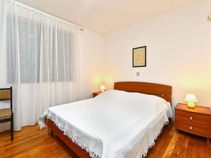Een opgemaakt bed met een witte deken, een houten nachtkastje met een lamp, een ingelijst beeld aan de muur en een raam met dunne gordijnen.