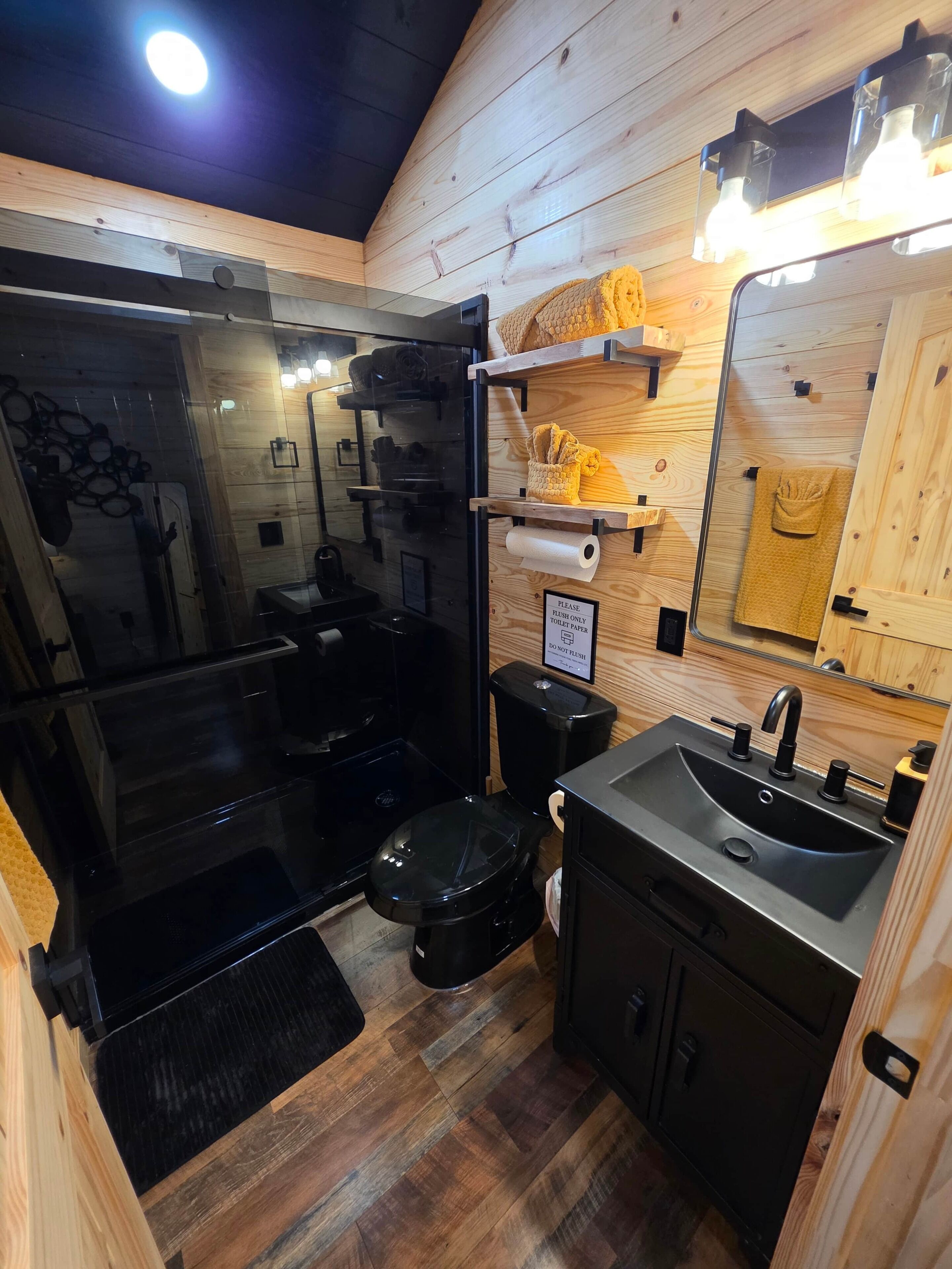 Un baño pequeño con cabina de ducha de vidrio, inodoro negro, lavabo con espejo y estantes de madera con toallas.