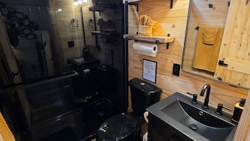 Un baño pequeño con cabina de ducha de vidrio, inodoro negro, lavabo con espejo y estantes de madera con toallas.