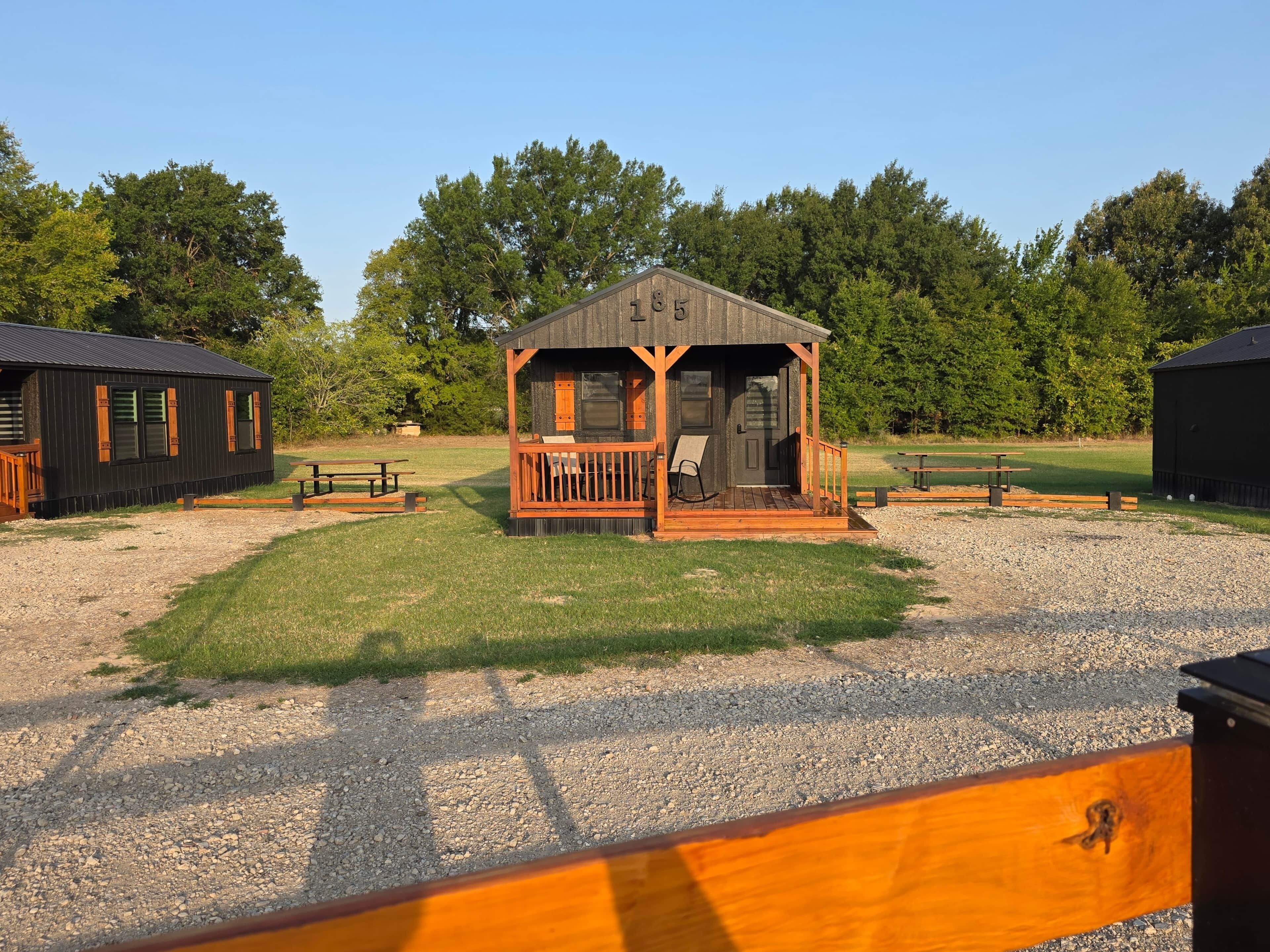 A small wooden cabin with a porch, surrounded by a gravel area and trees.