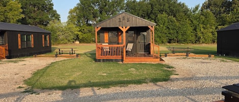 A small wooden cabin with a porch, surrounded by a gravel area and trees.
