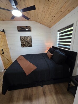 Ein Schlafzimmer mit einem Bett, einem Deckenventilator, einem Fenster mit Jalousien, einem an der Wand befestigten Kunstwerk und einem Holzschrank.