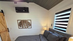 Un dormitorio con cama, lámparas de noche, ventana con persianas, conducto de aire en la pared y armario de madera.