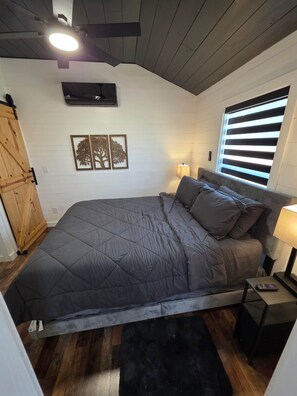 Ein Schlafzimmer mit einem Bett, Nachttischlampen, einem Fenster mit Jalousien, einem Wandlüfter und einem Holzschrank.