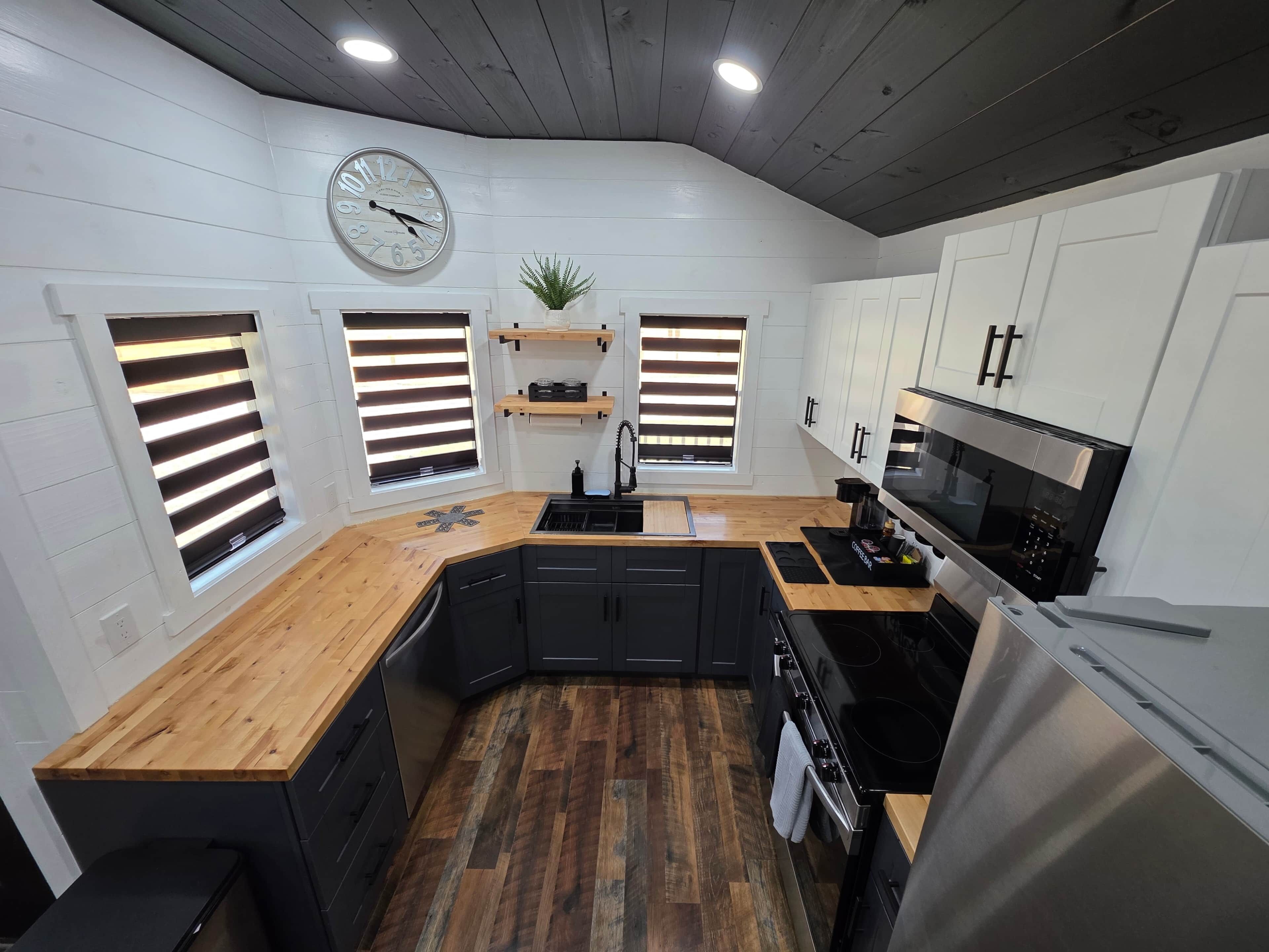 A compact kitchen with a wooden countertop, black cabinets, a stainless steel stove, and a clock on the wall.