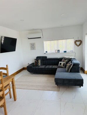 Uma sala de estar com sofá seccional, uma cadeira de madeira, televisão de tela plana na parede, janela com persianas e um coração decorativo na parede.