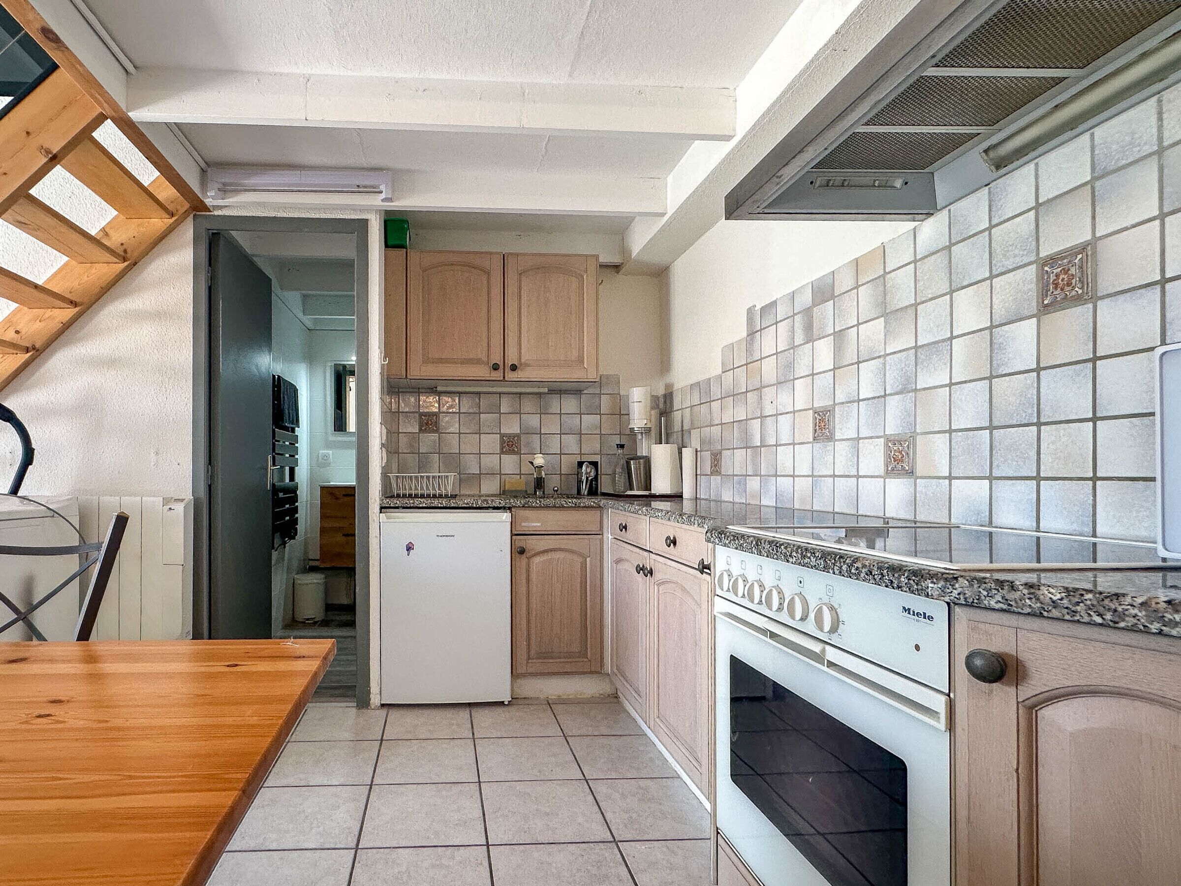 Una cocina con armarios de madera, salpicadero azulejado, horno empotrado y un comedor con mesa de madera.
