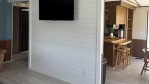 A modern interior with a flat-screen TV mounted on a white wall, a kitchen area with wooden cabinets, and a dining space with stools. - The River View at Esther’s (Vevay)