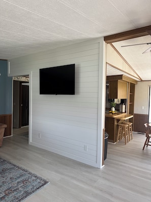 A modern interior with a flat-screen TV mounted on a white wall, a kitchen area with wooden cabinets, and a dining space with stools. - The River View at Esther’s (Vevay)