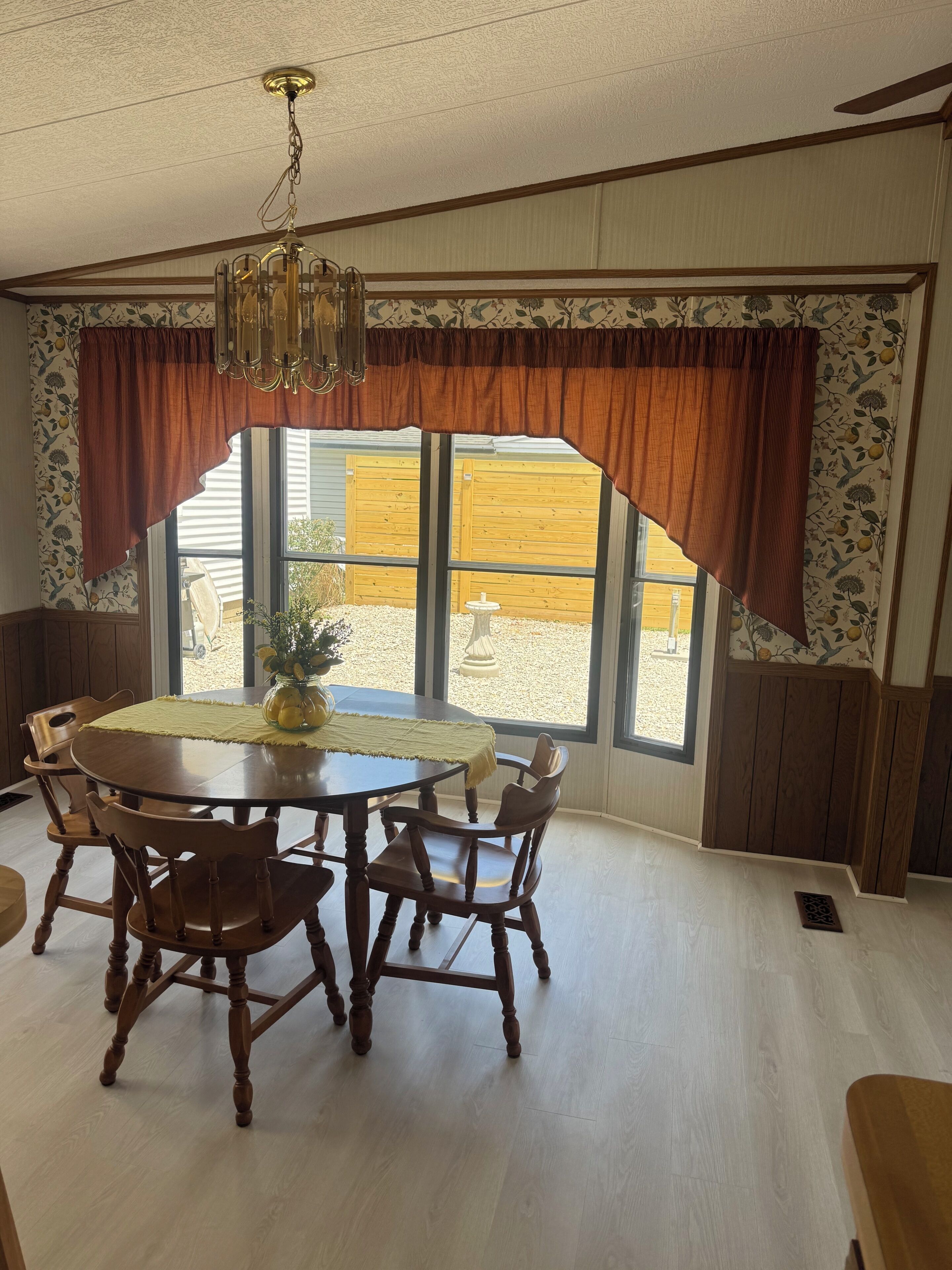A dining room with a round wooden table, four chairs, a chandelier, and large windows with curtains.
