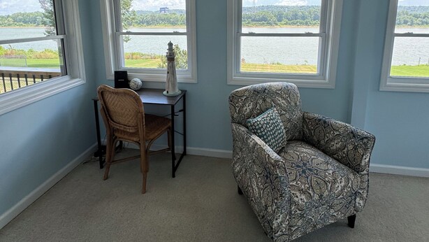 A room with a view of a lake, a patterned armchair, a wooden chair, and a small table with a lamp. - The River View at Esther’s (Vevay)