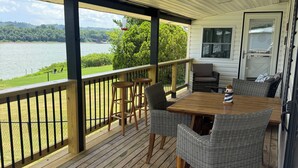 A covered deck with a wooden table and wicker chairs, overlooking a lake. - The River View at Esther’s (Vevay)