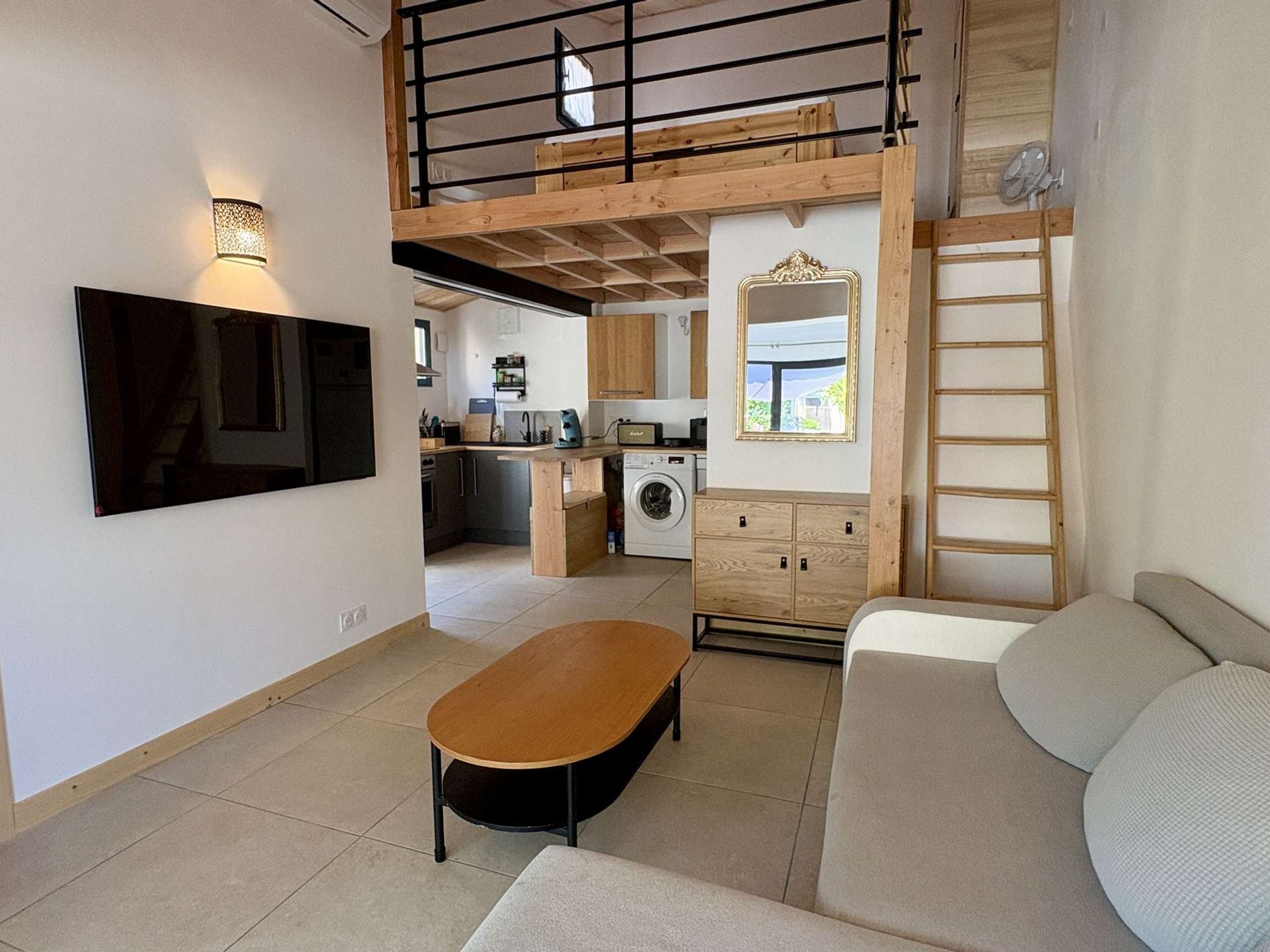 A compact living space with a kitchenette, a sofa, a wooden loft, and a television mounted on the wall.