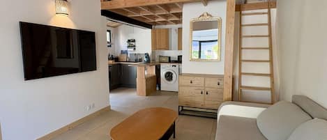 A compact living space with a kitchenette, a sofa, a wooden loft, and a television mounted on the wall.