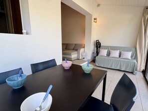 Ein Essbereich mit Tisch, Stühlen und Schüsseln mit Besteck. Im Hintergrund befindet sich eine Couch und ein Ventilator.