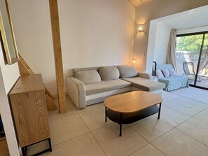 Un salon moderne avec un canapé d’angle, une table basse en bois et une vue sur l’extérieur grâce à une porte vitrée.
