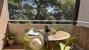 A round wooden table with green chairs, a woven hat, and a small potted plant on a balcony with a view of trees. - Apartment with pool, parking - 6 guests (Saint-Aygulf)