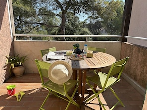 A round wooden table with green chairs, a woven hat, and a small potted plant on a balcony with a view of trees. - Apartment with pool, parking - 6 guests (Saint-Aygulf)