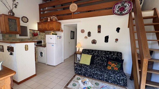 A compact living space with a kitchenette, a sofa with patterned cushions, a small table with children's books and toys, and a wooden ladder. - Apartment with pool, parking - 6 guests (Saint-Aygulf)