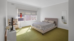 Quarto com cama, mesa de cabeceira, espelho, janela com persianas e sapateira.