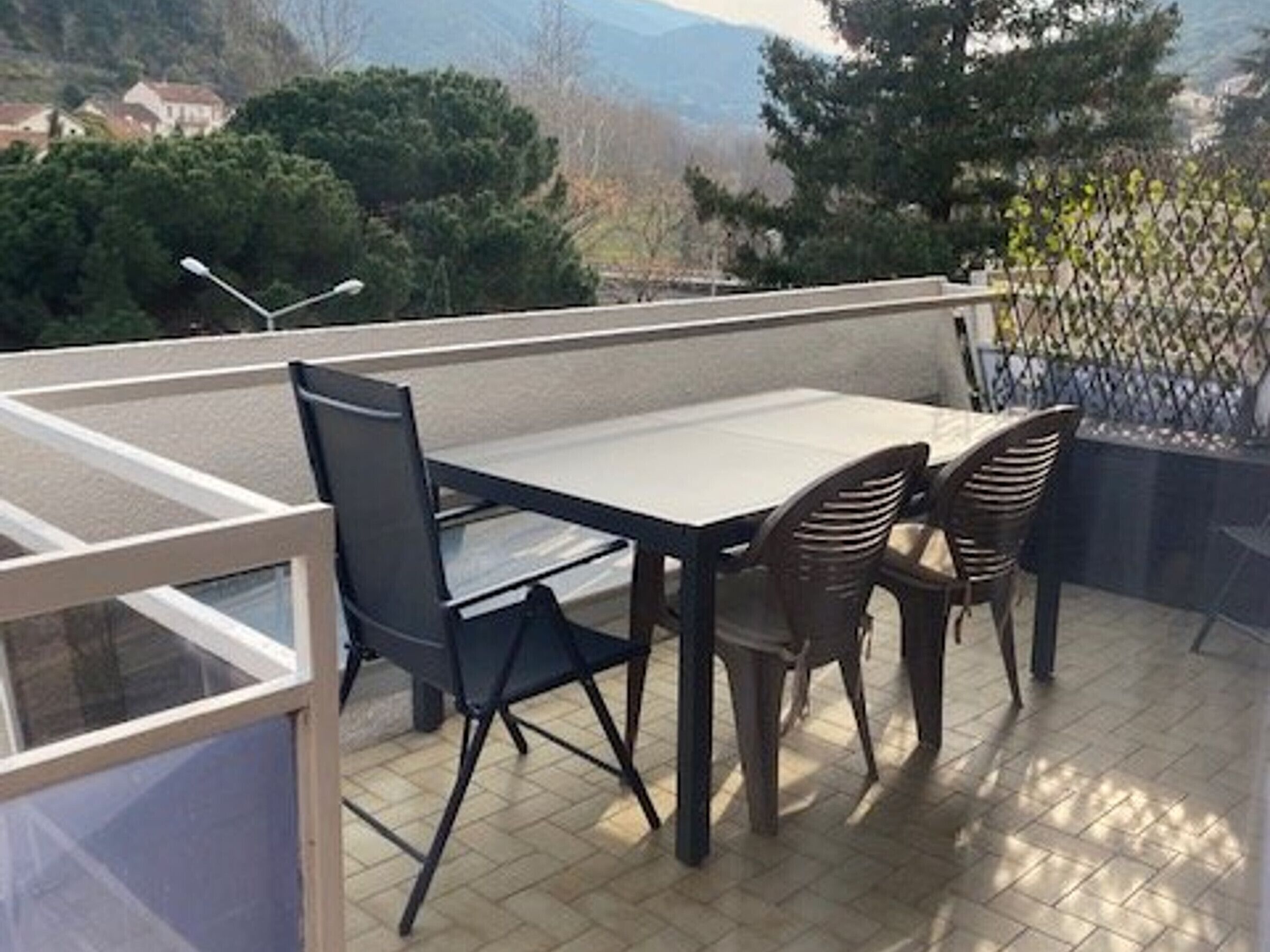 Een balkon met een tafel en stoelen, uitzicht op bomen en huizen, en een metalen hek.