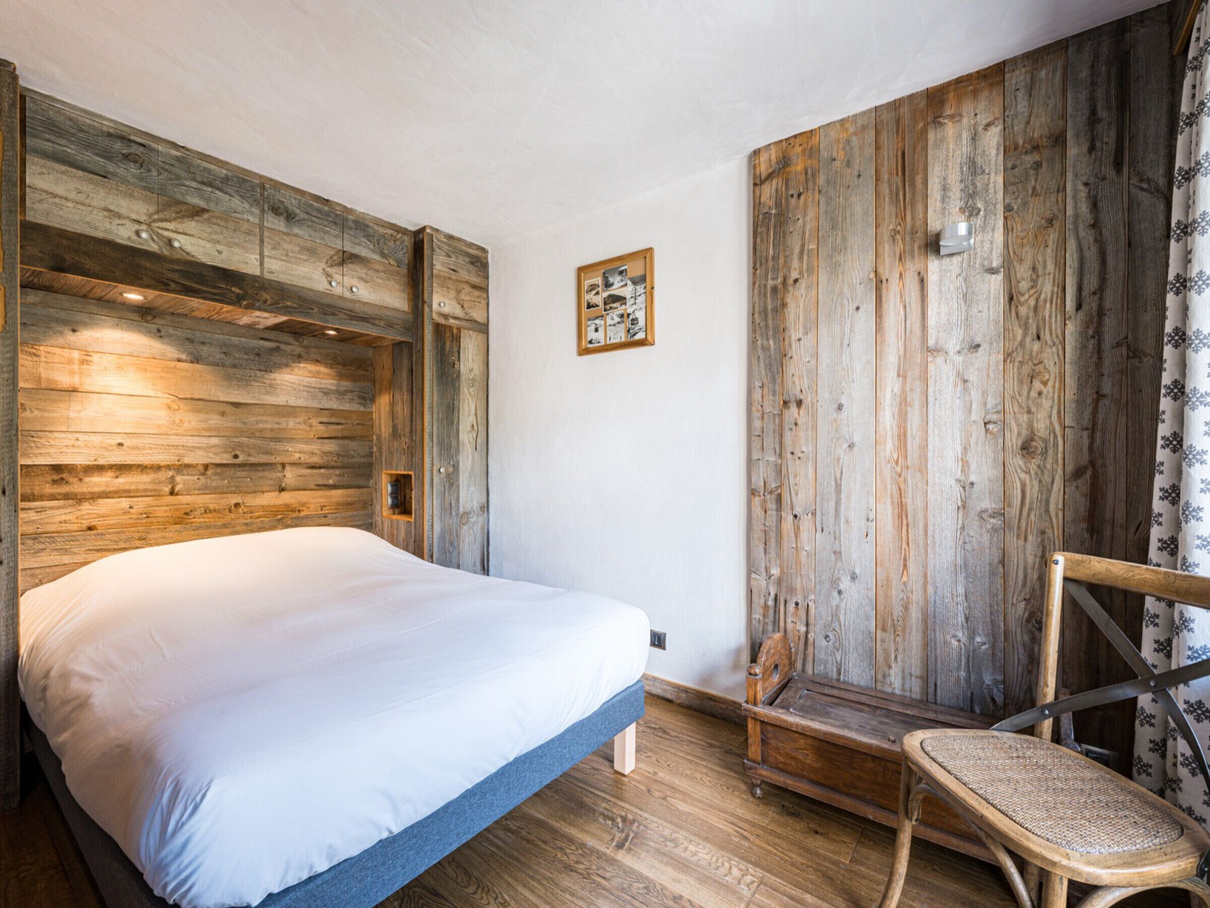 A room with a bed, wooden walls, a small wooden chair, and a picture on the wall.