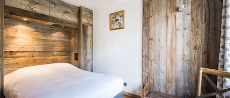A room with a bed, wooden walls, a small wooden chair, and a picture on the wall.
